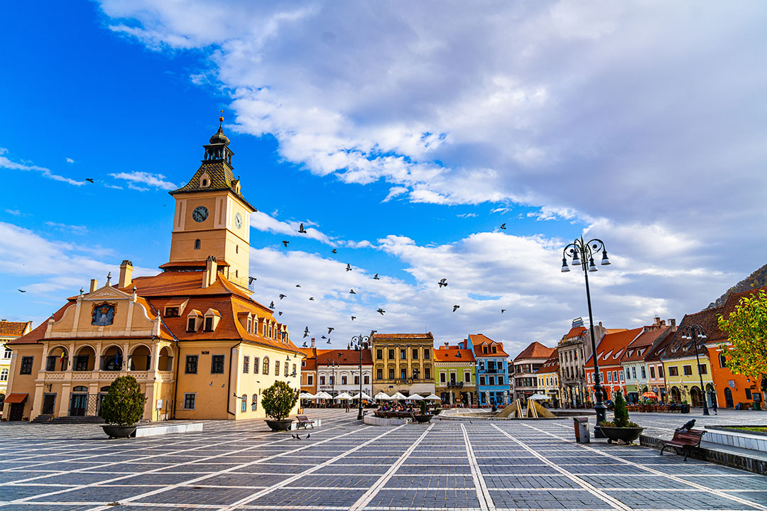 Colorful buildings in Brașov square. A vibrant square in Brasov, Romania features diverse architecture, a clock tower, and birds in flight against a clear blue sky.